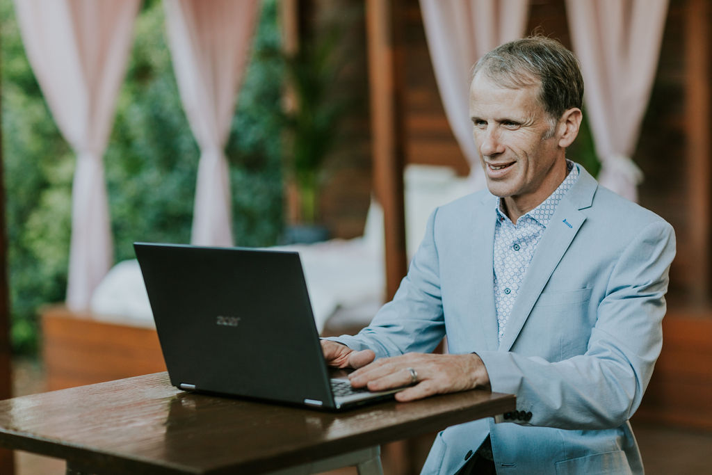 Loding your NOIM form via video call with legendary Elopement Celebrant Jamie Eastgate pictured in a stylish pale blue blazer on a laptop speaking with a couple on video call about their wedding and witnessing the couple sign their Notice Of Intention to Marry (NOIM) form.